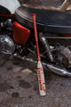 2027 Combat MFG Spec A1 Steel Horses Red Rider -3 BBCOR Baseball Bat resting against a motorcycle